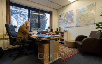 Persoon aan het werk in kantoorruimte huren in Almelo, gelegen op Bedrijvenpark Twente 305, met bureau, stoelen, kaarten aan de muur en raam met tuinzicht.