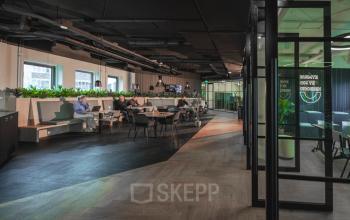 Moderne kantoorruimte huren op Egbert Gorterstraat 2-14 in Almelo met flexibele werkplekken, planten en glazen afscheidingen. Mensen werken en zitten aan tafels in een open vloerplan omgeving.