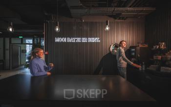 Twee mensen in een moderne kantoorruimte op Egbert Gorterstraat 2-14, Almelo, genieten van een koffiepauze. Industrieel interieur met een opvallende lichtinstallatie. Ideaal om te huren voor een creatieve werkomgeving.