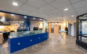 Moderne kantoorruimte met een receptiebalie, verlichte muurpanelen en open werkplekken in Plesmanweg 9, Almelo. Personen zijn actief bezig met zakelijke taken. Ideaal voor kantoorruimte huren.