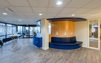 Moderne, open kantoorruimte op Plesmanweg 9, Almelo met houten vloer, blauwe zithoek, en grote ramen. Ideaal voor kantoorruimte huren en zakelijke ontmoetingen.