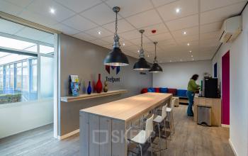 Kantoorruimte te huur op Plesmanweg 9, Almelo; interieur met houten tafel, witte stoelen, decoratieve vazen en persoon bezig bij koffiemachine.