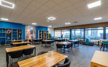 Kantoorruimte huren op Plesmanweg 9 in Almelo met een ruime en moderne kantine, voorzien van houten tafels, zwarte stoelen en decoratieve elementen. Een persoon zit aan tafel met een laptop.