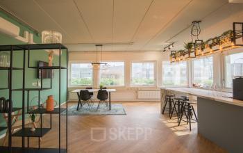 Licht, modern kantoorpand met houten vloer en grote ramen in Almere Poort aan Kabelstraat 5. Ruimte is ingericht als gezamenlijke werkplek met planten en stoelen rondom houten tafels.