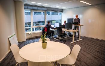 Moderne kantoorruimte huren op P.J. Oudweg 41 in Almere Stad, met verstelbare bureaus, een tafel met stoelen en uitzicht naar buiten. Personeel werkt aan bureaus.