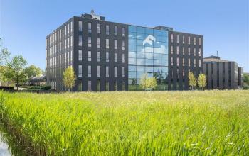 Modern kantoorpand aan de Flemingweg 8 in Alphen aan den Rijn, te huur via Sollf. Gebouw met zwarte gevel en grote ramen, omringd door groene vegetatie.