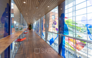 Moderne gang met glazen wanden en kleurrijke decoraties in kantoorpand op Stationsplein 49, Amersfoort Centraal Station. Hoge barkrukken langs houten tafel bieden uitzicht op buiten.