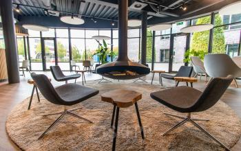 Moderne lounge met grijze stoelen en houten tafels in het kantoorpand op Databankweg 26, Amersfoort De Hoef. Grote ramen bieden veel natuurlijk licht.