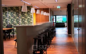 Gezellige kantoorruimte huren op Prof. W.H. Keesomlaan 12 in Amstelveen met lange bar, houten vloer en groene muurdecoratie. Moderne inrichting met hoge krukken en vergaderruimtes.