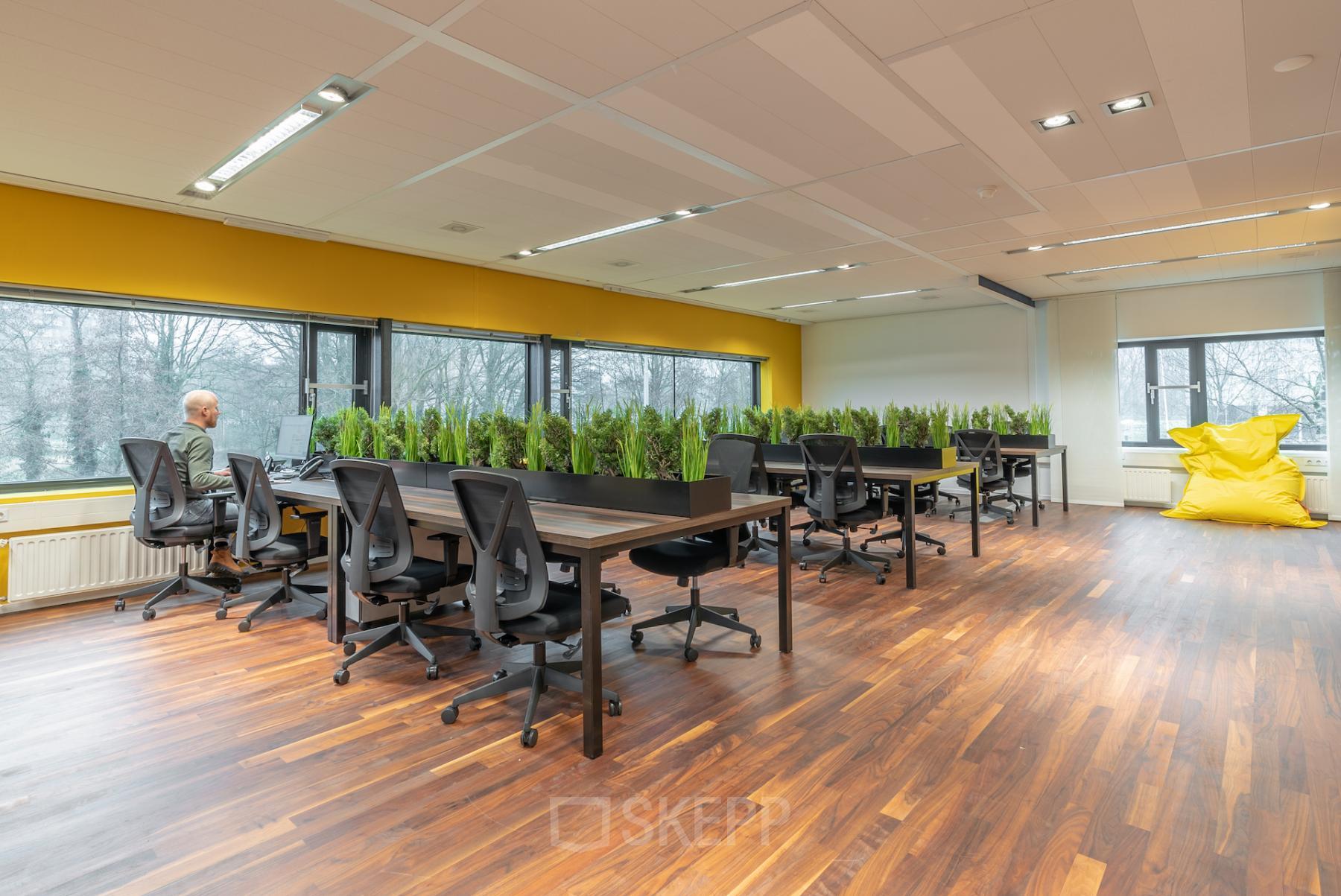 Kantoorruimte te huur op Prof. W.H. Keesomlaan 12, Amstelveen. Moderne werkruimte met houten vloeren, planten als scheidingswanden en veel lichtinval. Enkele mensen zijn aan het werk aan bureaus.