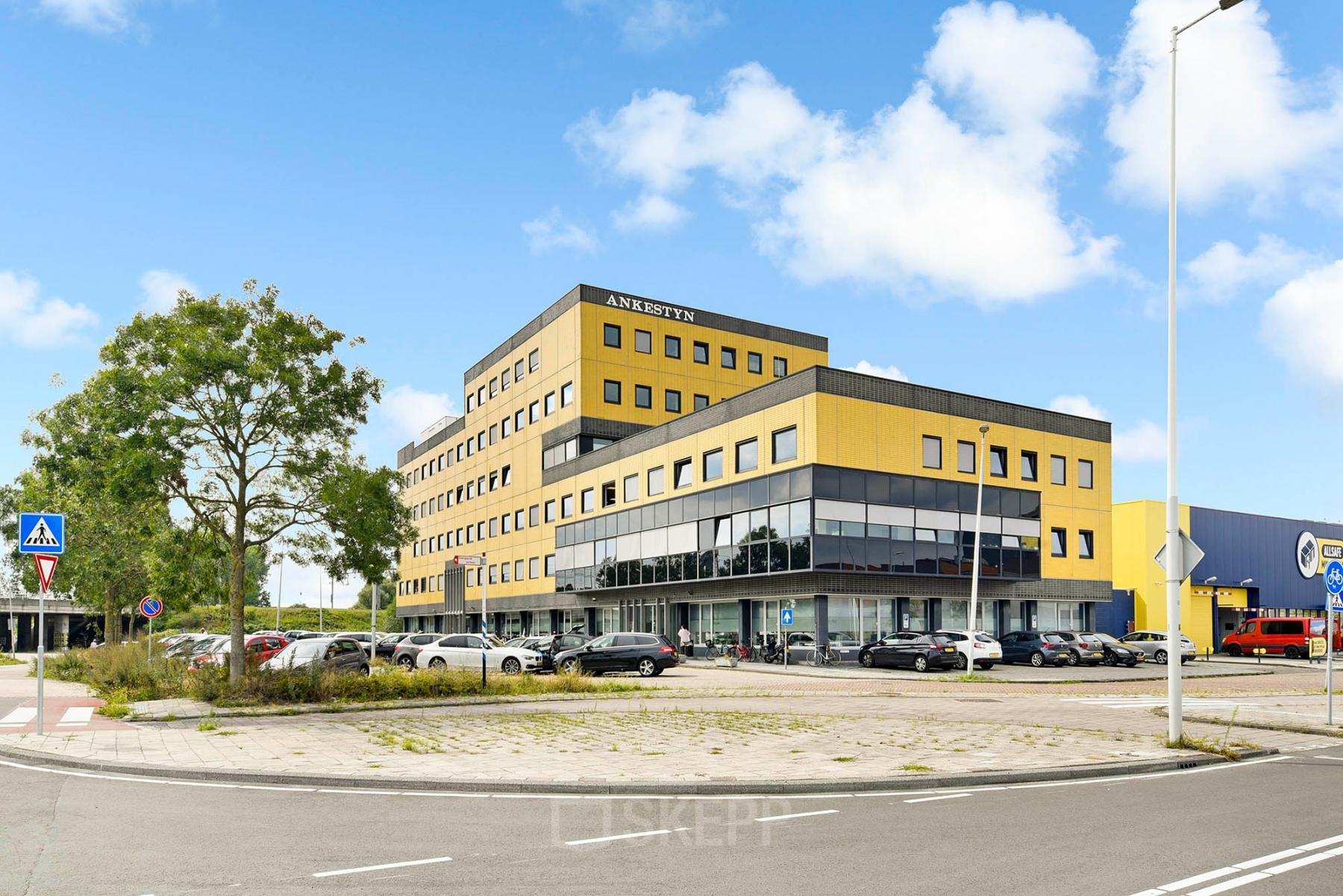 Exterieur van kantoorpand op Joan Muyskenweg 82, gelegen in Amsterdam Amstel Business Park, met opvallende gele facade en parkeerplaats. Perfecte locatie om kantoorruimte te huren voor ondernemingen.