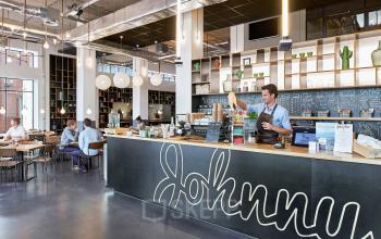 Moderne lounge en werkruimte in kantoorpand gelegen aan Joan Muyskenweg 22, Amsterdam Amstel Business Park. Medewerker serveert koffie aan de balie. Mensen zitten aan tafels in gesprek.
