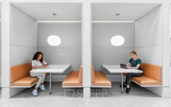 Twee personen werken aan aparte tafels in moderne kantoorruimte met grijze wanden en oranje zitbanken in Mr. Treublaan 7, Amsterdam Amstel.