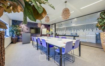 Moderne vergaderruimte in een kantoorpand op Prins Bernhardplein 200, Amsterdam Amstel, met een lange tafel en paarse stoelen, omgeven door planten en glazen wanden.