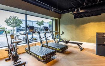 Kantoorpand op Zwaansvliet 1 in Amsterdam Buitenveldert met fitnessapparatuur, waaronder loopbanden en een roeimachine, bij grote ramen met uitzicht op de parkeerplaats.