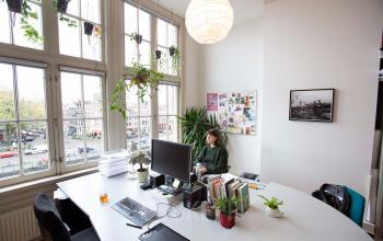 Kantoorruimte huren aan de Schippersgracht 1-3, Amsterdam Centrum, met grote ramen en veel natuurlijke lichtinval. Een persoon werkt aan een bureau met computer en kantoorbenodigdheden.