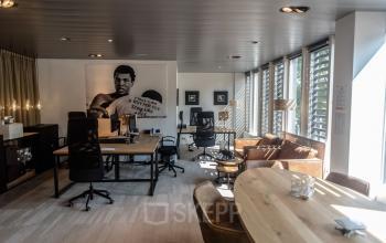 Kantoorruimte huren op Muiderstraat 1, Amsterdam Centrum. Moderne inrichting met grote ramen en meerdere bureaus. Vergaderruimte in gebruik, terwijl mensen aan het werk zijn. Zonnig en goed verlicht interieur.