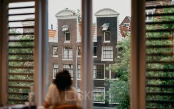 Uitzicht vanuit een kantoor op de Muiderstraat 1 in Amsterdam Centrum, met een persoon die aan een bureau zit en werkt. Historische grachtenpanden zijn zichtbaar door het raam.