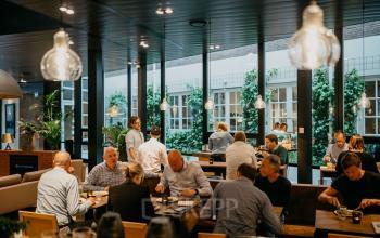 Kantoorruimte in Amsterdam Centrum op Muiderstraat 1. Mensen zijn bezig met zakelijke lunches en besprekingen in een gemeubileerde eetgelegenheid met grote ramen en veel groen.
