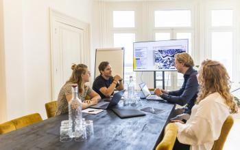 Zakelijke meeting in een kantoor op Prins Hendrikkade 21, Amsterdam Centrum. Collega's overleggen rond een tafel met laptops en een scherm aan de muur.