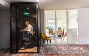 Moderne kantoorruimte huren op Prins Hendrikkade 21, Amsterdam Centrum met een flexibele werkplek voor individueel gebruik. Een persoon werkt geconcentreerd in een afgeschermde cabine, terwijl andere werkplekken beschikbaar zijn.