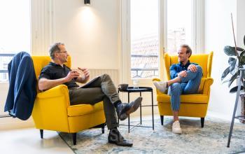 Twee personen zitten in een stijlvolle kantoorruimte aan de Singel 126 in Amsterdam Centrum, bezig met een zakelijke bespreking. Gele fauteuils en een salontafel sieren de ruimte.