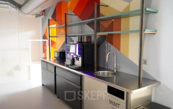 Kantoorruimte met kleurrijke wandkunst, een moderne koffiemachine en gootsteen, gelegen op Weesperplein 4b in Amsterdam Centrum. Geschikt voor koffie- en pauzeruimte in een kantoorpand te huur.