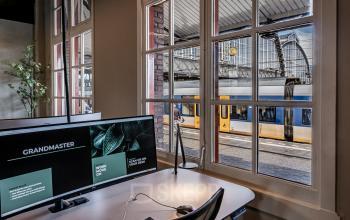Kantoorruimte huren op Stationsplein 9, Amsterdam Centrum met uitzicht op het station door grote raampartijen, gemeubileerd met een bureau en monitor.
