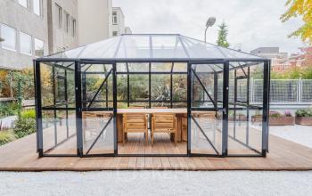Glazen paviljoen bij Weesperstraat 107-121 in Amsterdam Centrum, met houten tafels en stoelen, ideaal voor creatieve ontmoetingen of alternatieve werkruimtes. Perfecte locatie voor kantoorruimte huren.