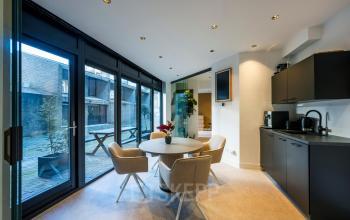 Moderne kantoorruimte huren gelegen aan Groenmarktkade 5-H in Amsterdam Centrum met stijlvolle afwerking, voorzien van een ronde tafel, beige stoelen, kitchenette en grote ramen met uitzicht op een binnenplaats.