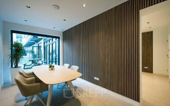 Moderne kantoorruimte huren in Amsterdam Centrum met grote glazen pui, houten wandpanelen en vier comfortabele stoelen rond een tafel. Groenmarktkade 5-H biedt een stijlvolle werkplek.