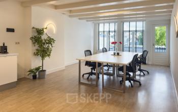 Ruime kantoorruimte huren aan Warmoesstraat 155 in Amsterdam Centrum met grote ramen, houten vloer en moderne bureaus. De zonnige werkplek is ingericht met comfortabele stoelen en groene accenten, ideaal voor productiviteit.