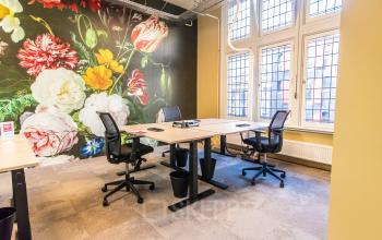 Lichte kantoorruimte te huur op Nieuwezijds Voorburgwal 104-108 in Amsterdam Centrum, met kleurrijke bloemenmuur, bureaus en stoelen. Grote ramen bieden veel daglicht.