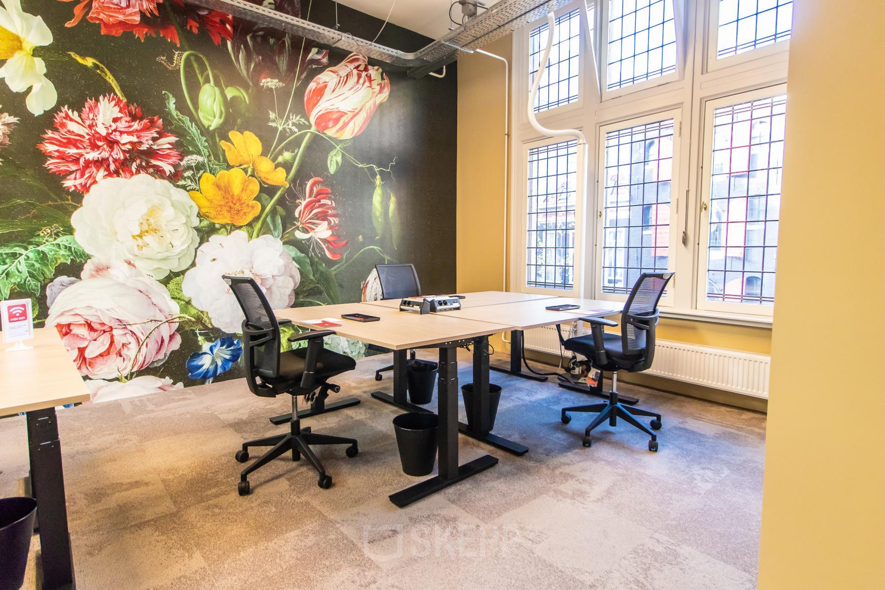 Lichte kantoorruimte te huur op Nieuwezijds Voorburgwal 104-108 in Amsterdam Centrum, met kleurrijke bloemenmuur, bureaus en stoelen. Grote ramen bieden veel daglicht.