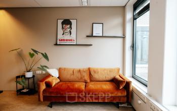 Gezellige kantoorruimte met een oranje bank tegen een muur met planken en een plant, perfect om te huren in Amsterdam De Schinkel.