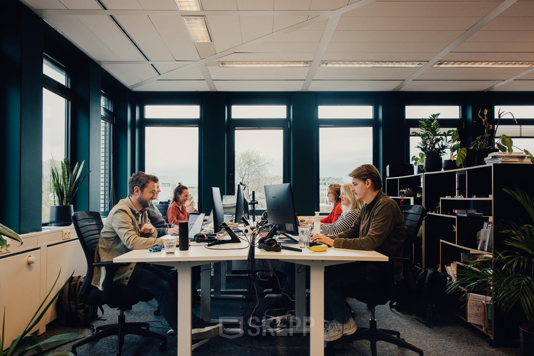 Zakelijke kantoorruimte te huur op IJsbaanpad 2 in Amsterdam De Schinkel, met moderne werkplekken en groene accenten. Mensen aan het werk aan bureaus met computers.