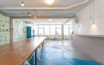 Kantoorruimte met lange houten tafels en stoelen, grote ramen, veel natuurlijk licht, en hanglampen aan het plafond op Generaal Vetterstraat 77e, Amsterdam De Schinkel.