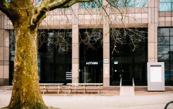 Kantoorgebouw aan de Overschiestraat 182 in Amsterdam De Schinkel met grote glazen gevel en entree met houten picknicktafels ervoor. Ideale locatie voor kantoorruimte huren in een moderne omgeving.