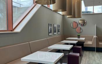 Moderne kantoorruimte met gezellige zitplekken en tafels. Grote lampen hangen aan het plafond en de ruimte heeft een open uitstraling met veel natuurlijk licht.