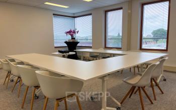 Vergaderruimte in Amsterdam Duivendrecht met moderne witte stoelen en tafels in U-vorm, grote ramen en decoratieve bloemen. Perfect voor het huren van kantoorruimte voor meetings of presentaties.
