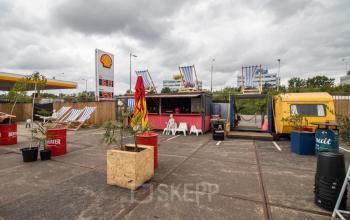 Exterieur van een kantoorpand op Anthony Fokkerweg 3, Amsterdam Nieuw-West, met een Shell-tankstation op de achtergrond, inclusief kleurrijke zitplaatsen en een foodtruck, wat een creatieve werkomgeving impliceert.