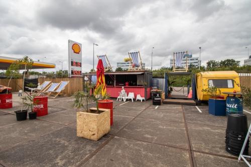 Exterieur van een kantoorpand op Anthony Fokkerweg 3, Amsterdam Nieuw-West, met een Shell-tankstation op de achtergrond, inclusief kleurrijke zitplaatsen en een foodtruck, wat een creatieve werkomgeving impliceert.