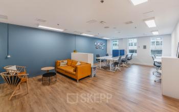 Ruime kantoorpand op Koningin Wilhelminaplein 1, Amsterdam Nieuw-West, met houten vloer, moderne werkplekken, een oranje bank en blauwe muren. Perfect voor wie kantoorruimte huren overweegt.