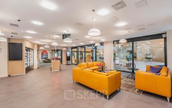 Grote lounge met gele banken, glazen wanden en diverse verlichting in kantoorpand aan Koningin Wilhelminaplein 1, ideaal voor kantoorruimte huren in Amsterdam Nieuw-West.