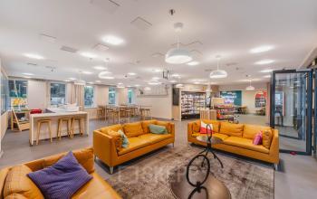 Lichte en ruime lounge in een kantoorpand op Koningin Wilhelminaplein 1, Amsterdam Nieuw-West, met comfortabele gele banken en moderne verlichting. Ideaal voor ontspanning en informele bijeenkomsten. Perfecte plek voor kantoorruimte huren.