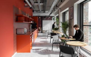 Moderne kantoorruimte huren in Amsterdam Noord met felrode keukenwand, houten tafels, en stoelen. Personen werken op laptops aan tafels bij grote ramen met veel natuurlijk licht.