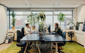 Kantoorruimte huren op TT Vasumweg 44 in Amsterdam Noord met uitzicht en veel groen. Twee mensen voeren zakelijke activiteiten uit aan hun bureaus.