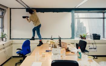 Een kantoor in Amsterdam Noord, TT Vasumweg 44, met een persoon die een muur schildert en bureaus die laptops, kantoorspullen en planten bevatten.