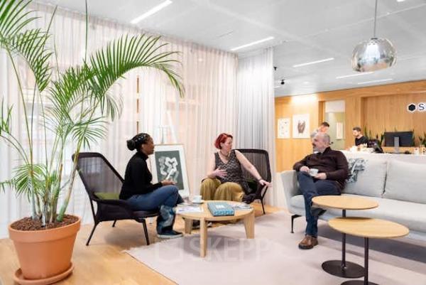 Drie mensen in een ontspannen gesprek in een modern kantoorpand in Amsterdam Noord, gelegen aan Mt. Lincolnweg 38-40. De ruimte is stijlvol ingericht met comfortabele stoelen en een grote plant.