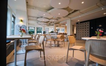 Moderne, lichte kantine met houten stoelen en tafels in kantoorpand op Apollolaan 151, Amsterdam Oud Zuid. Grote ramen zorgen voor veel daglicht, ideaal voor huur van kantoorruimte met een frisse uitstraling.